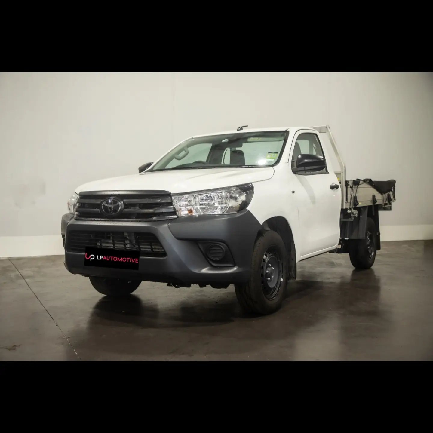 A white Toyota Hilux cab-chassis photographed against the white wall at LP Automotive, wearing an LP Automotive dealer plate